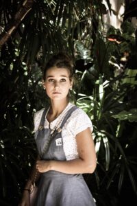 A young woman with tied-back hair and a serious expression stands amidst lush tropical foliage. She is wearing a white shirt with small patterns and a denim-style pinafore dress, holding a small purse with a chain strap. Sunlight filters through the leaves, creating dappled shadows.