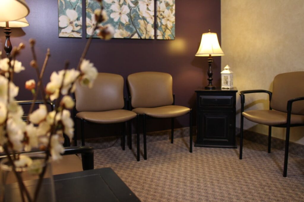 A cozy waiting room with three beige chairs against a dark purple wall. There is a small black side table with a lamp and decorative lantern. The floor is carpeted, and a vase with white flowers is blurred in the foreground.