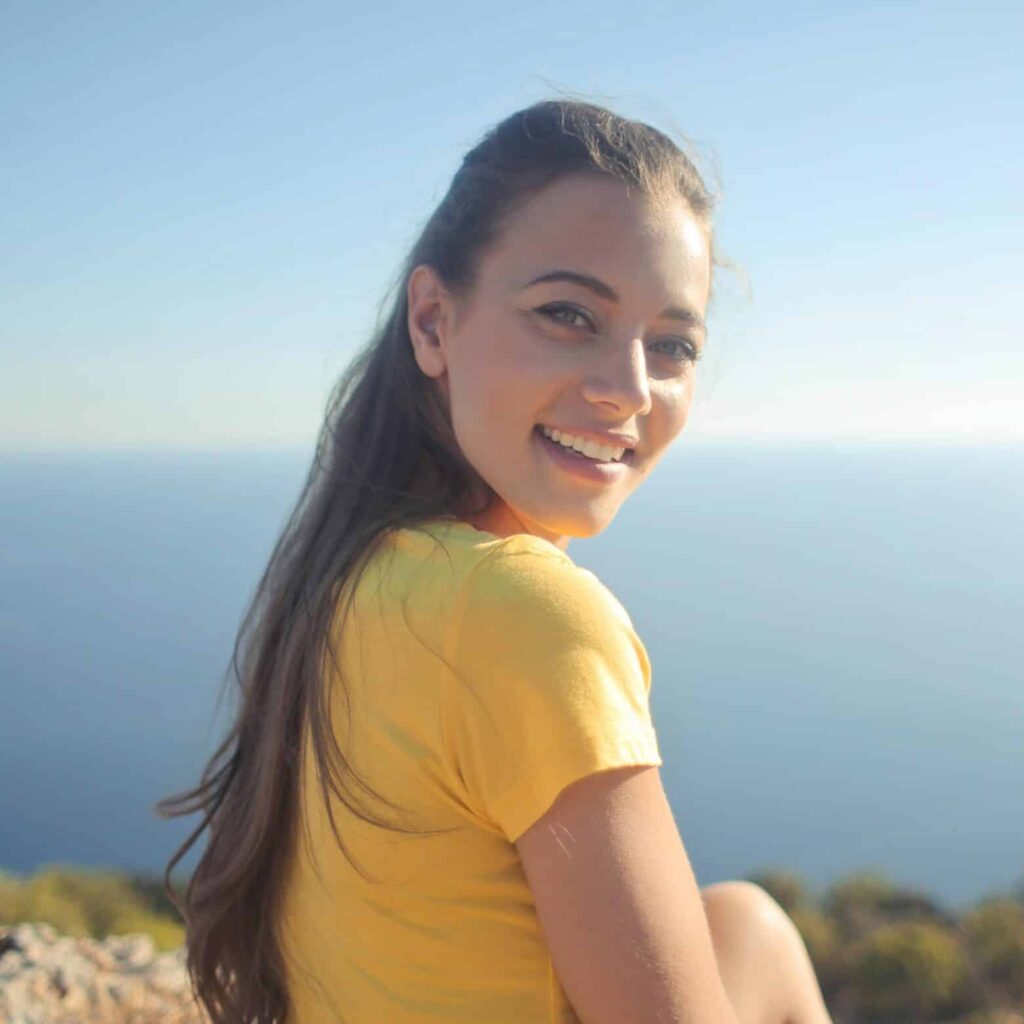 A woman with long hair is smiling while sitting on a cliffside with a clear blue ocean and sky in the background. She is wearing a yellow shirt, and the sunlight highlights her face, giving a warm and cheerful impression. The landscape suggests a serene and scenic location.