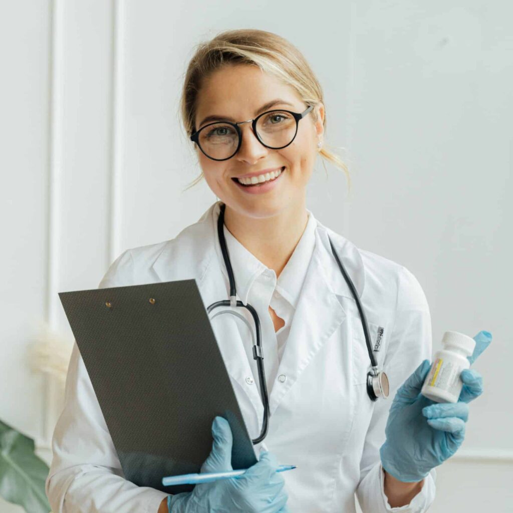 A female doctor wearing glasses, a white coat, and a stethoscope, holding a clipboard and a prescription bottle. She is smiling and wearing blue gloves, standing against a plain white background.