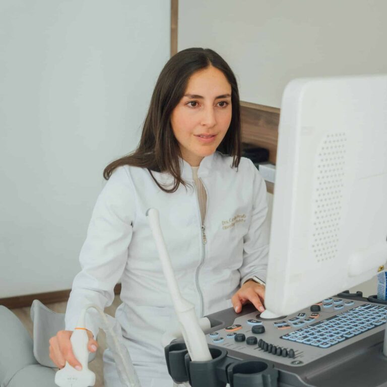 A woman in a white medical coat is using an ultrasound machine. She is holding a probe and looking at a monitor, indicating she is performing a medical ultrasound exam.