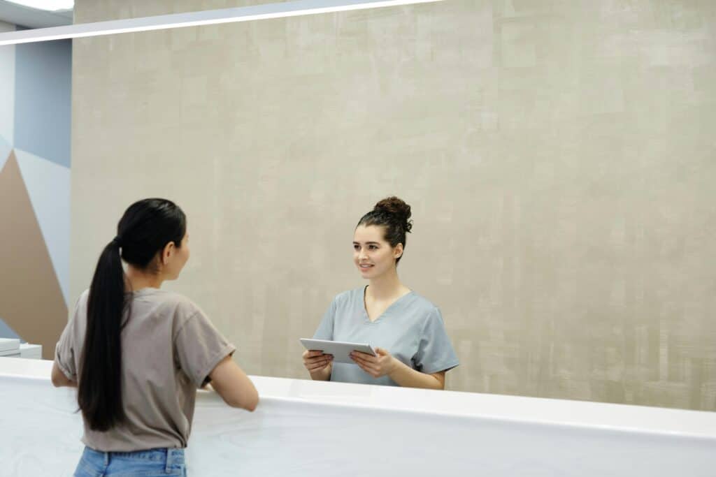 A receptionist in medical scrubs holding a tablet and smiling at a woman with long hair at a reception desk in a modern office setting.