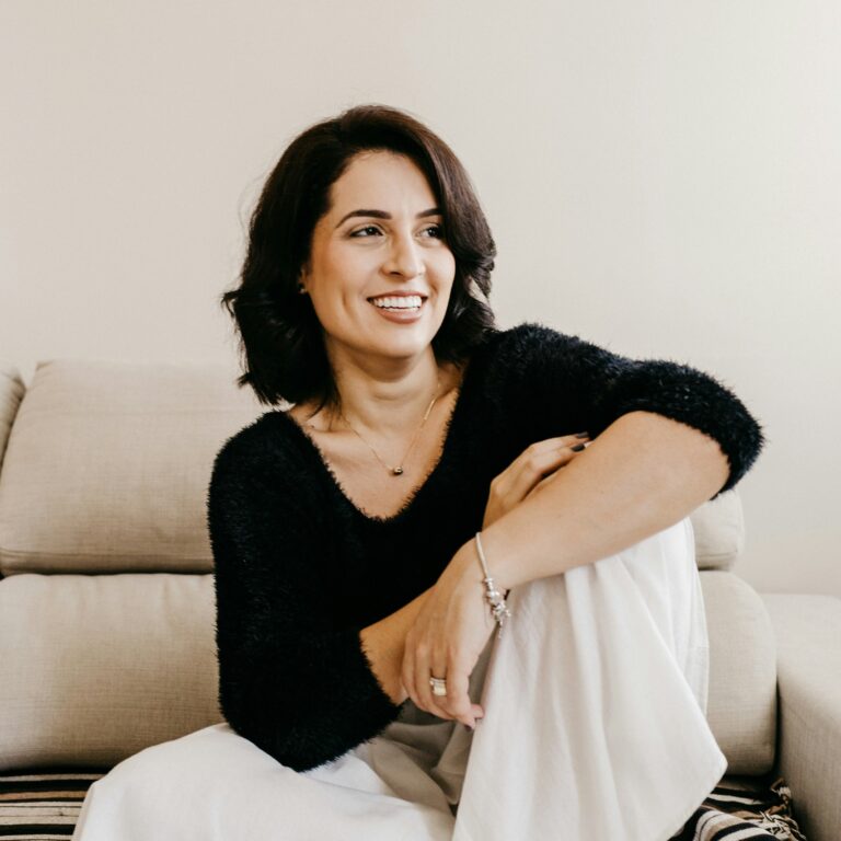 A woman with dark hair smiling while sitting on a beige couch. She is wearing a black fuzzy sweater and white pants, with one arm resting on her knee. The background is a plain light-colored wall.
